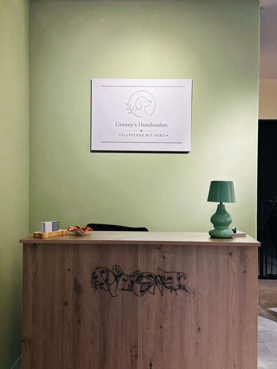 Empfangstresen aus Holz mit Hundegesichtern aus Draht, darüber das Logo-Schild von Greeny's Hundesalon an einer salbeigrünen Wand, daneben eine grüne Tischlampe