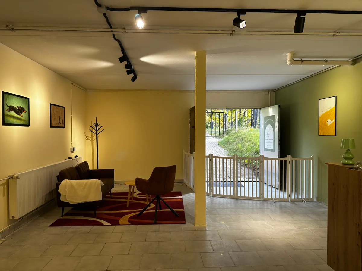 Wartebereich mit Sofa, Sessel und Teppich, Hundebilder an den Wänden; durch die offene Tür der Weg nach draußen zum Tor